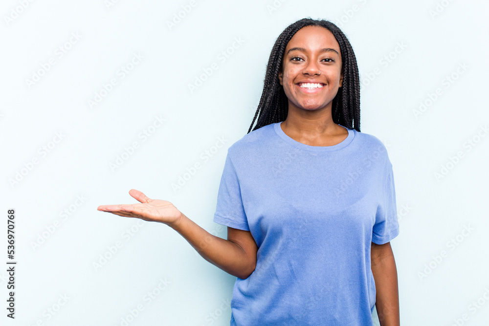 Young African American woman with braids hair isolated on blue background showing a copy space on a palm and holding another hand on waist. Young African American woman with braids hair isolated on blue background showing a copy space on a palm and holding another hand on waist.
