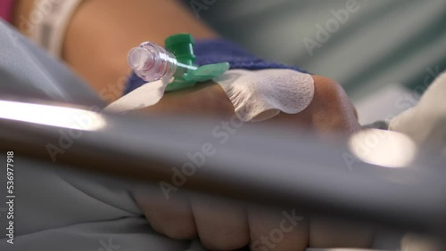 Close-up of a Caucasian arm with drip in hospital bed clutching the sheet.