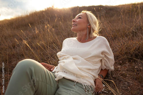 A Mature woman dressed boho style clothis posing in the field with grass against warm light of sun