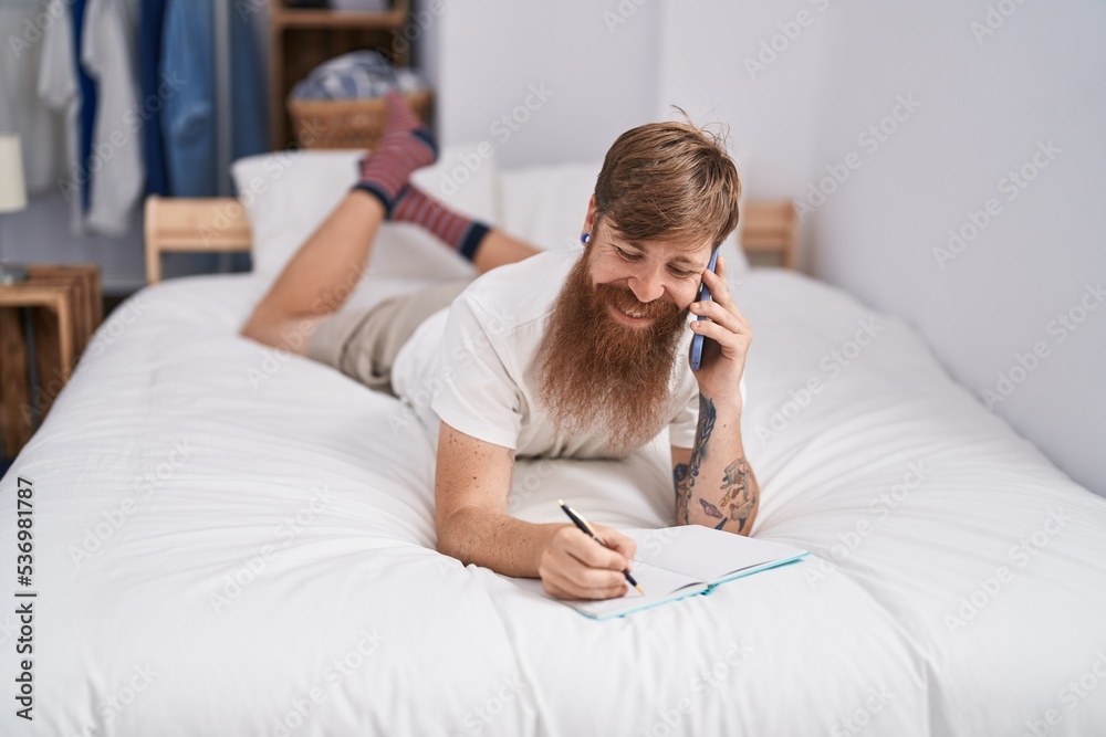 Young redhead man talking on smartphone writing on book at bedroom