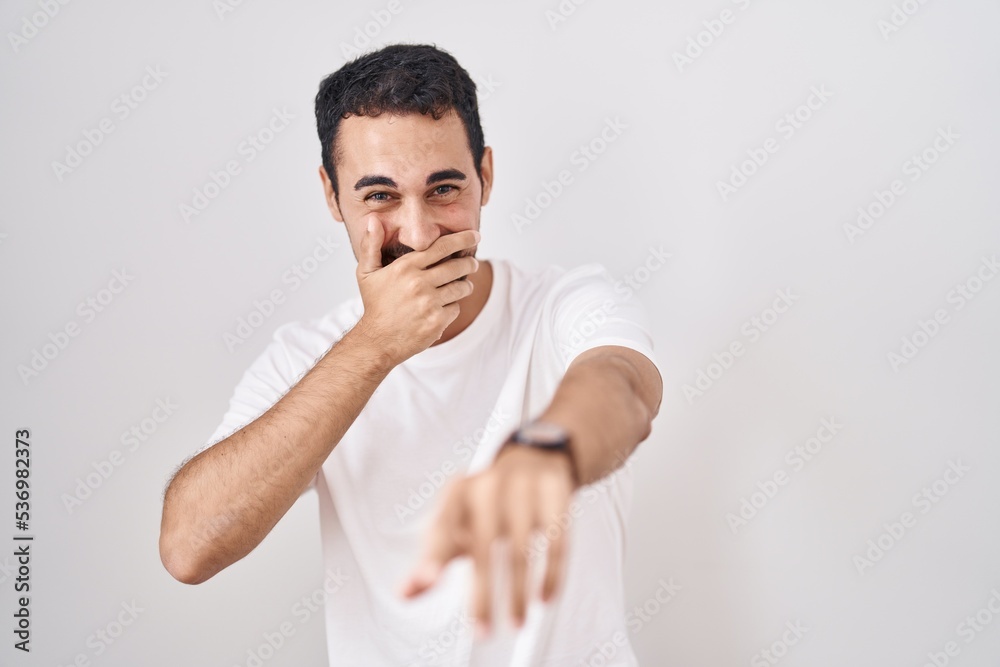 Handsome hispanic man standing over white background laughing at you, pointing finger to the camera with hand over mouth, shame expression Handsome hispanic man standing over white background laughing at you, pointing finger to the camera with hand over mouth, shame expression