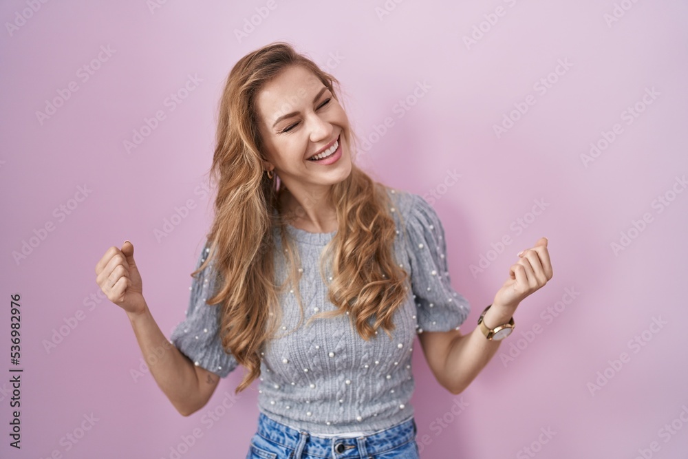 Obraz premium Beautiful blonde woman standing over pink background very happy and excited doing winner gesture with arms raised, smiling and screaming for success. celebration concept.