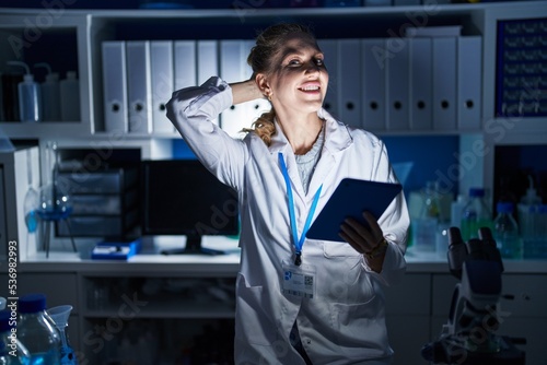 Beautiful blonde woman working at scientist laboratory late at night smiling confident touching hair with hand up gesture  posing attractive and fashionable