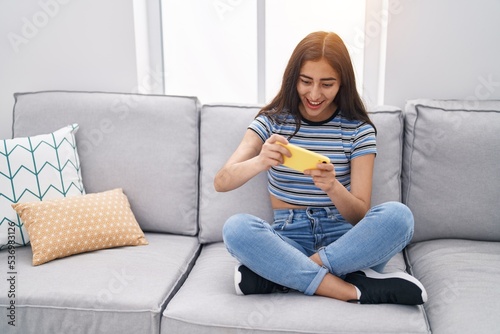 Young hispanic girl playing video game by smartphone at home