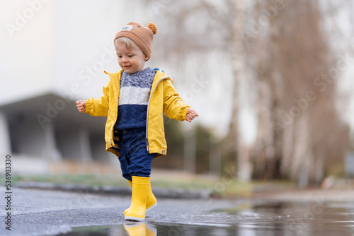 Happy little boy in yellow raincoat jumping in puddle after rain in autumn day.