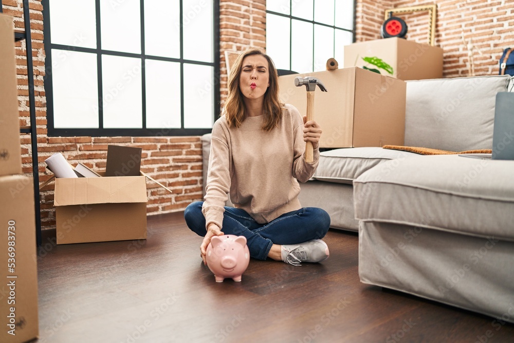 Young woman holding piggy bank and hammer moving to a new home making fish face with mouth and squinting eyes, crazy and comical. Young woman holding piggy bank and hammer moving to a new home making fish face with mouth and squinting eyes, crazy and comical.