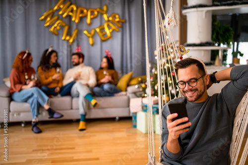 Friends having fun at a Christmas party in their decorated apartment, wearing festive sweaters, chatting and scrolling through their phone 