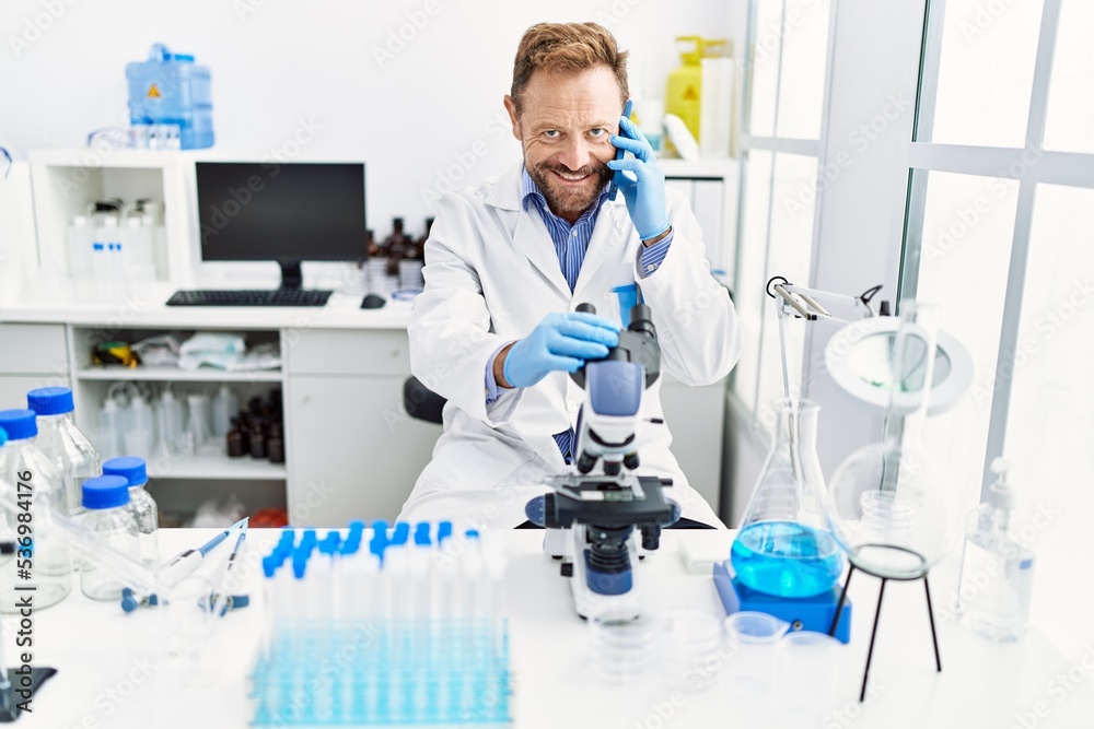 Fototapeta premium Middle age hispanic man wearing scientist uniform talking on the smartphone using microscope at laboratory