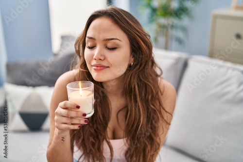 Young beautiful hispanic woman sitting on sofa smelling aromatic candle at home photo