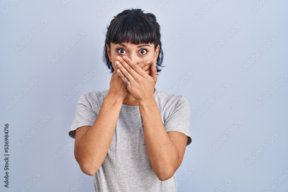 Young hispanic woman wearing casual t shirt over blue background shocked covering mouth with hands for mistake. secret concept. Young hispanic woman wearing casual t shirt over blue background shocked covering mouth with hands for mistake. secret concept.