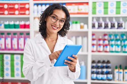 Young beautiful hispanic woman pharmacist using touchpad working at pharmacy