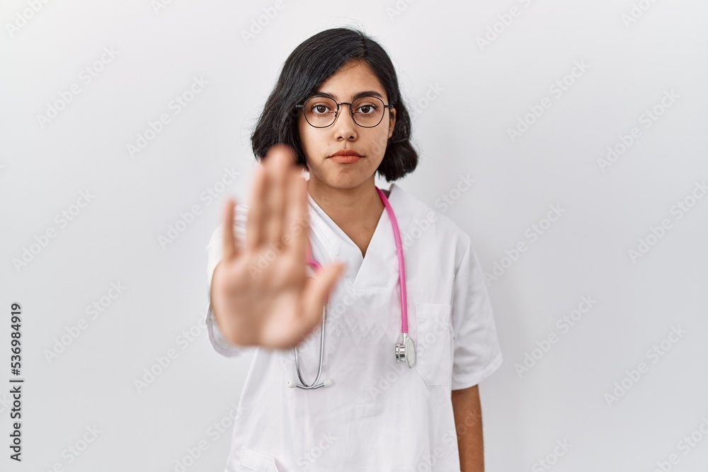 Young hispanic doctor woman wearing stethoscope over isolated background doing stop sing with palm of the hand. warning expression with negative and serious gesture on the face.
