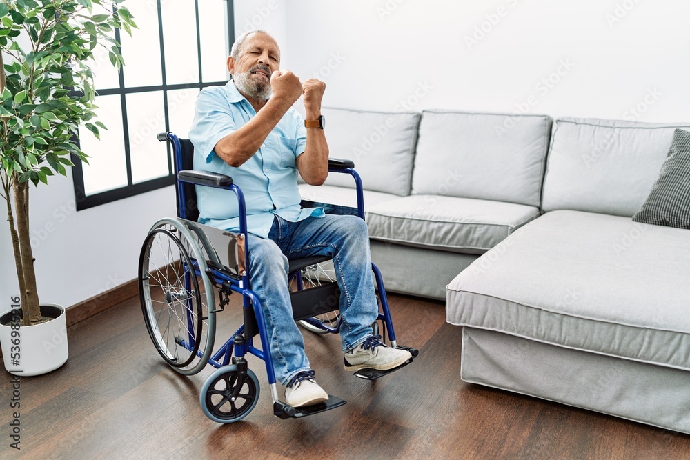 Handsome senior man sitting on wheelchair at the living room angry and mad raising fist frustrated and furious while shouting with anger. rage and aggressive concept. Handsome senior man sitting on wheelchair at the living room angry and mad raising fist frustrated and furious while shouting with anger. rage and aggressive concept.