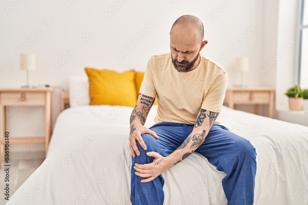 Young bald man suffering for knee injury sitting on bed at bedroom Young bald man suffering for knee injury sitting on bed at bedroom