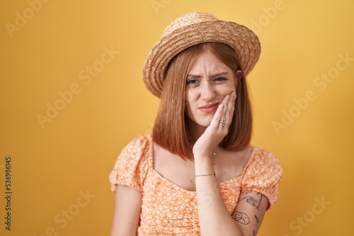 Young redhead woman standing over yellow background wearing summer hat touching mouth with hand with painful expression because of toothache or dental illness on teeth. dentist