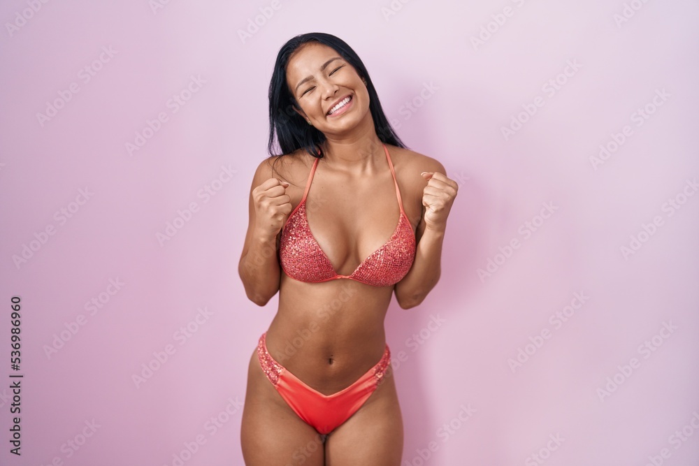 Hispanic woman wearing bikini excited for success with arms raised and eyes closed celebrating victory smiling. winner concept. Hispanic woman wearing bikini excited for success with arms raised and eyes closed celebrating victory smiling. winner concept.