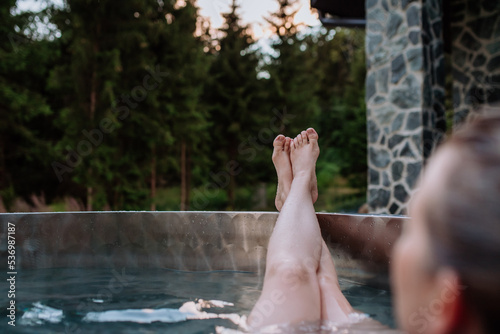 Unrecognizable young woman with feet up relaxing in hot tub outdoor in nature.