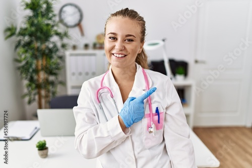 Young caucasian woman wearing doctor uniform and stethoscope at the clinic cheerful with a smile of face pointing with hand and finger up to the side with happy and natural expression on face