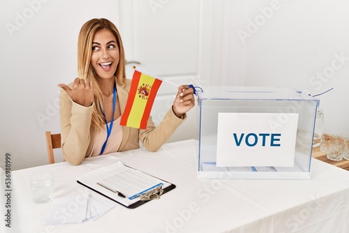 Blonde beautiful young woman at political campaign election holding spain flag pointing thumb up to the side smiling happy with open mouth