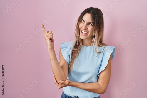 Young hispanic woman standing over pink background with a big smile on face, pointing with hand and finger to the side looking at the camera. © Krakenimages.com
