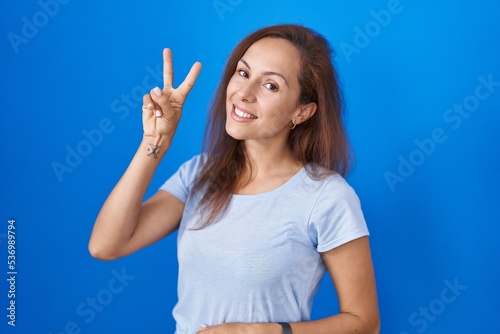 Brunette woman standing over blue background smiling looking to the camera showing fingers doing victory sign. number two.