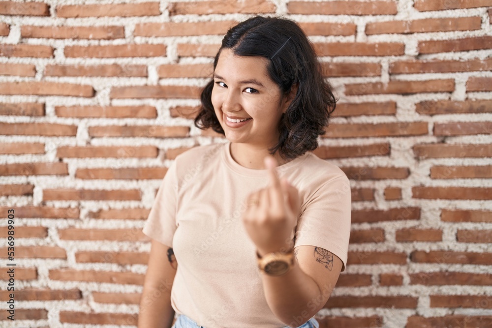 Young hispanic woman standing over bricks wall beckoning come here gesture with hand inviting welcoming happy and smiling Young hispanic woman standing over bricks wall beckoning come here gesture with hand inviting welcoming happy and smiling