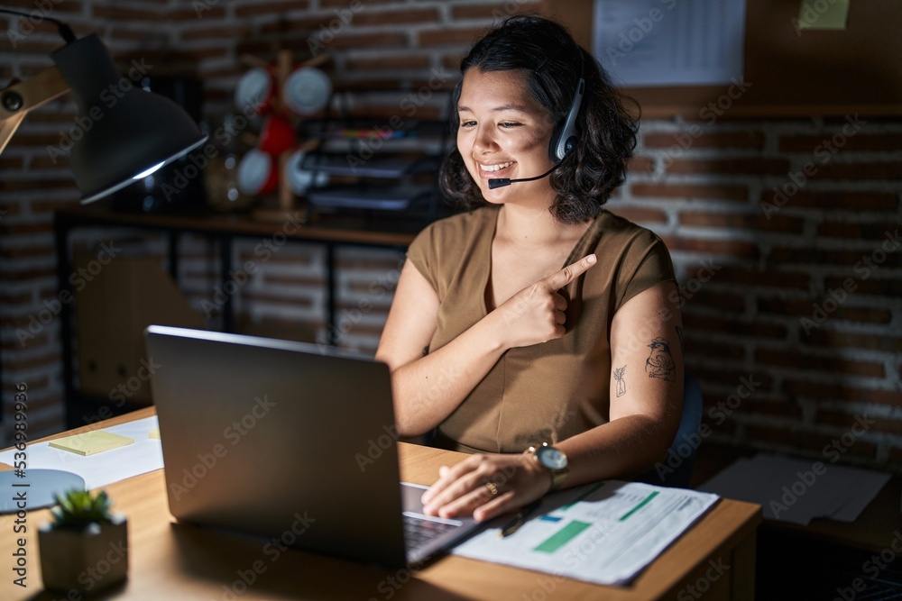 Young hispanic woman working at the office at night cheerful with a smile on face pointing with hand and finger up to the side with happy and natural expression Young hispanic woman working at the office at night cheerful with a smile on face pointing with hand and finger up to the side with happy and natural expression