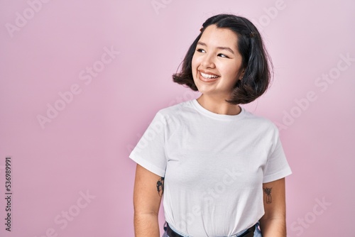 Young hispanic woman wearing casual white t shirt over pink background looking away to side with smile on face, natural expression. laughing confident.