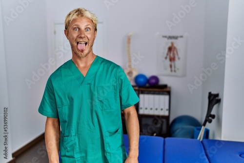 Young blond man wearing physiotherapist uniform standing at clinic sticking tongue out happy with funny expression. emotion concept.