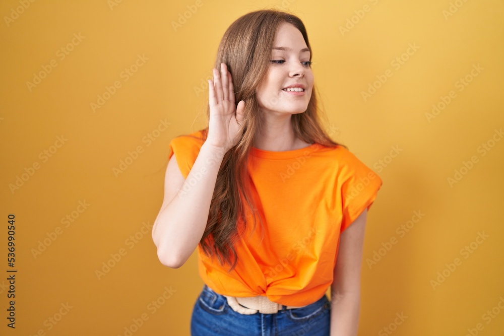 Caucasian woman standing over yellow background smiling with hand over ear listening an hearing to rumor or gossip. deafness concept. Caucasian woman standing over yellow background smiling with hand over ear listening an hearing to rumor or gossip. deafness concept.