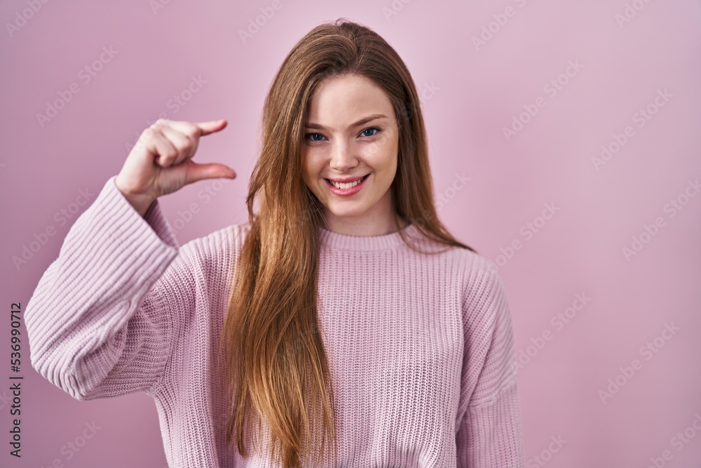 Young caucasian woman standing over pink background smiling and confident gesturing with hand doing small size sign with fingers looking and the camera. measure concept. Young caucasian woman standing over pink background smiling and confident gesturing with hand doing small size sign with fingers looking and the camera. measure concept.