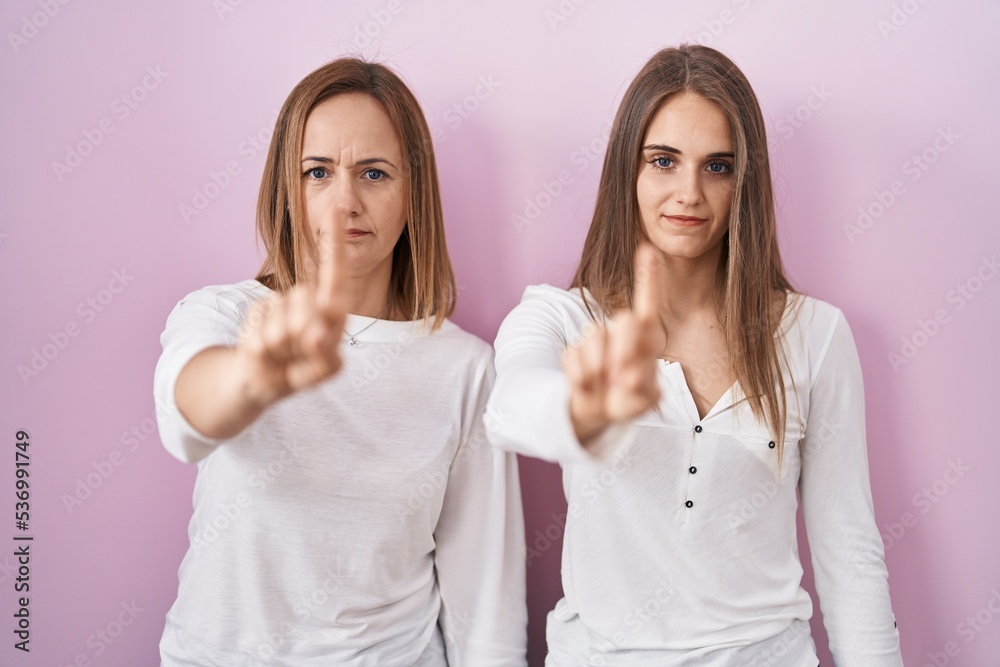 Middle age mother and young daughter standing over pink background pointing with finger up and angry expression, showing no gesture Middle age mother and young daughter standing over pink background pointing with finger up and angry expression, showing no gesture
