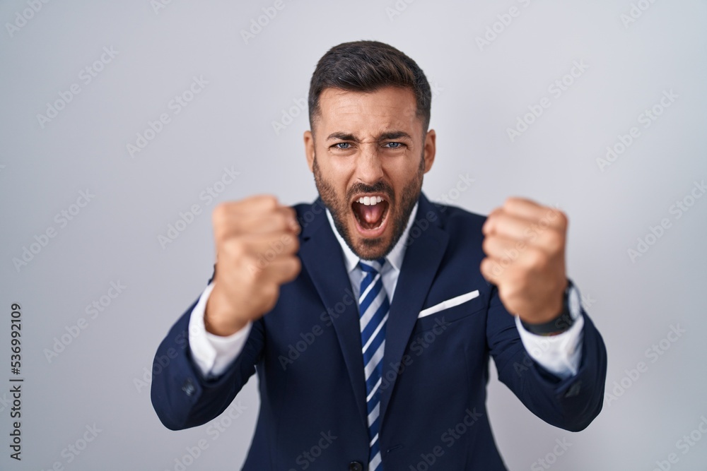 Handsome hispanic man wearing suit and tie angry and mad raising fists frustrated and furious while shouting with anger. rage and aggressive concept. Handsome hispanic man wearing suit and tie angry and mad raising fists frustrated and furious while shouting with anger. rage and aggressive concept.
