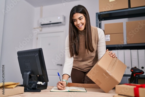 Young beautiful hispanic woman ecommerce business worker writing on notebook holding package at office