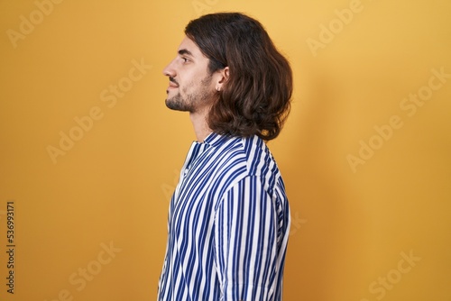 Hispanic man with long hair standing over yellow background looking to side, relax profile pose with natural face with confident smile.