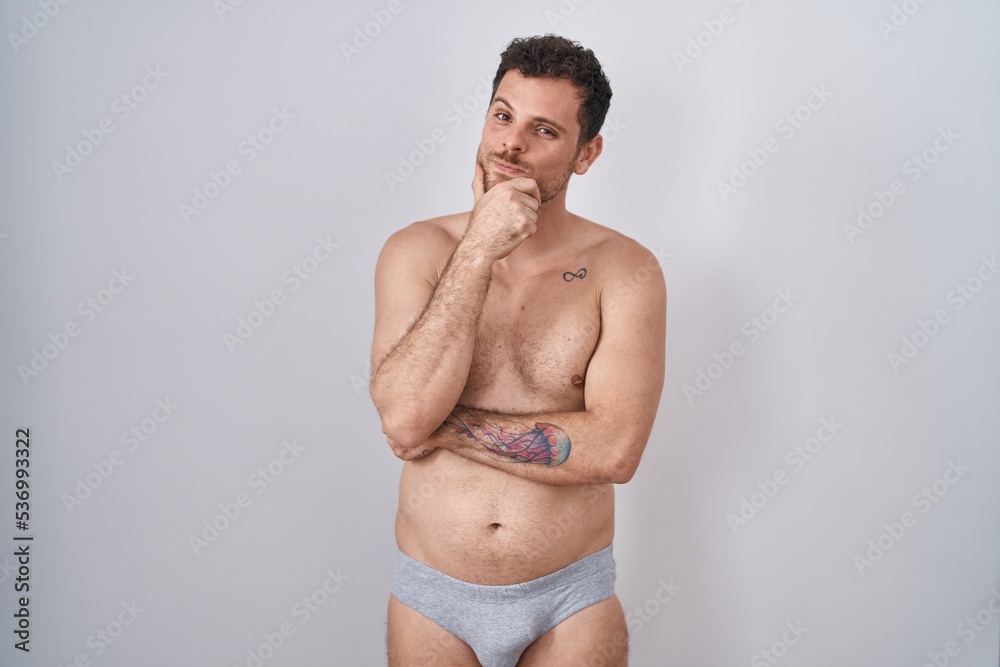 Fototapeta premium Young hispanic man standing shirtless wearing underware looking confident at the camera smiling with crossed arms and hand raised on chin. thinking positive.