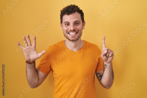Young hispanic man standing over yellow background showing and pointing up with fingers number six while smiling confident and happy.