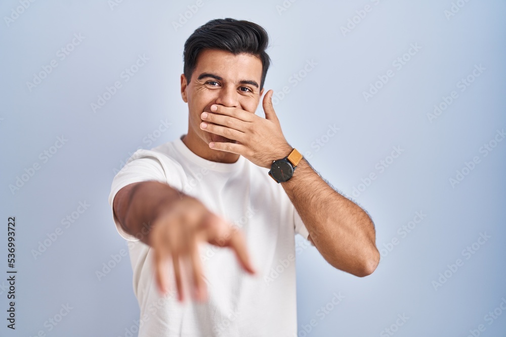 Hispanic man standing over blue background laughing at you, pointing finger to the camera with hand over mouth, shame expression Hispanic man standing over blue background laughing at you, pointing finger to the camera with hand over mouth, shame expression
