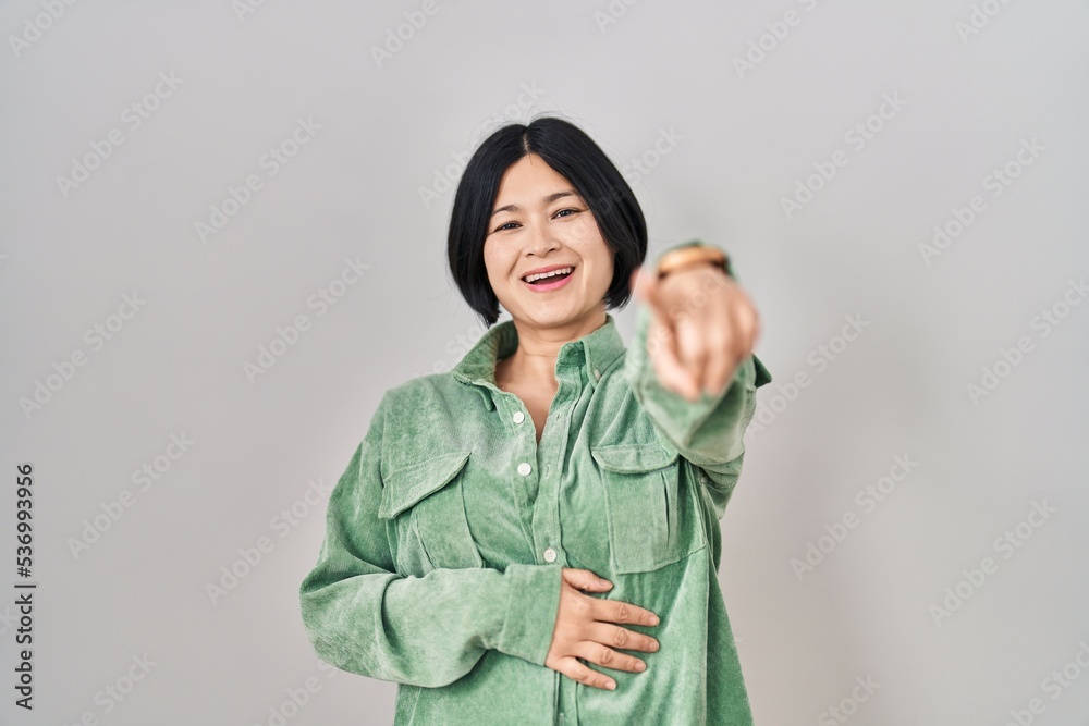 Young asian woman standing over white background laughing at you, pointing finger to the camera with hand over body, shame expression