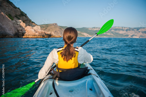 A girl is sailing in a kayak on the sea against the backdrop of mountains, rear view