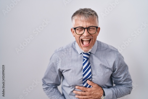 Hispanic business man with grey hair wearing glasses smiling and laughing hard out loud because funny crazy joke with hands on body.