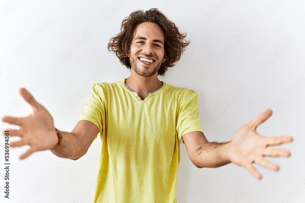 Young hispanic man standing over isolated background looking at the camera smiling with open arms for hug. cheerful expression embracing happiness.