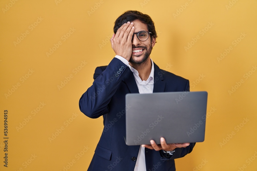 Handsome latin man working using computer laptop covering one eye with hand, confident smile on face and surprise emotion.