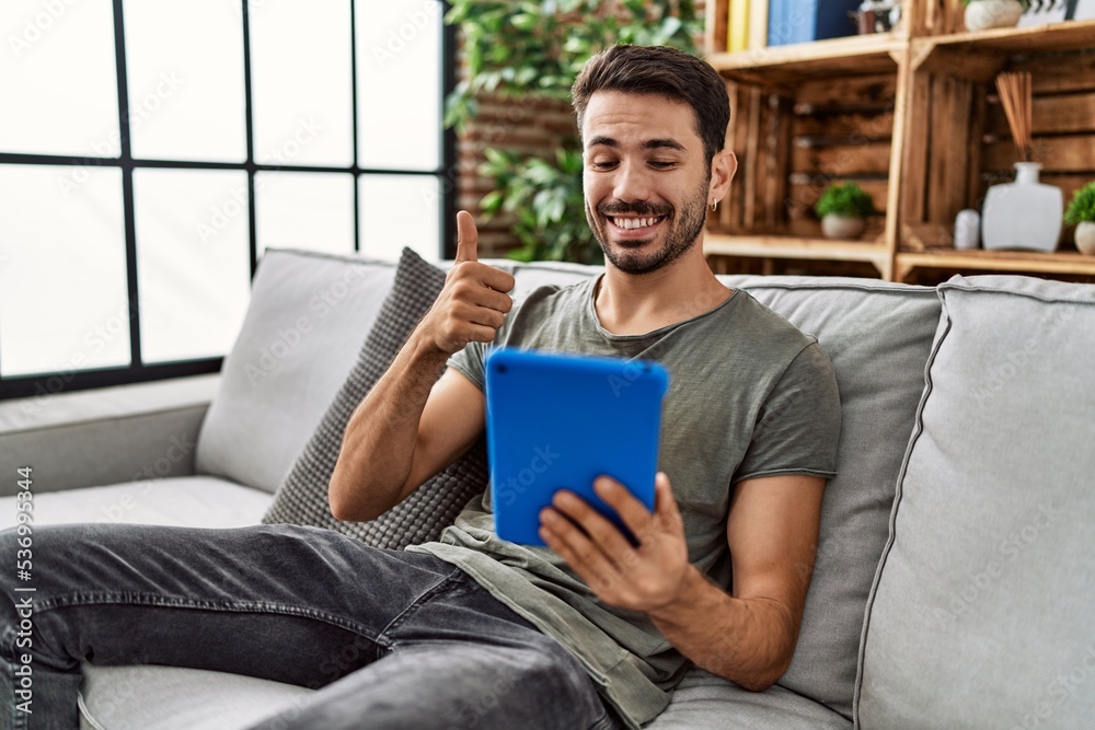 Young hispanic man using touchpad sitting on the sofa smiling happy and positive, thumb up doing excellent and approval sign