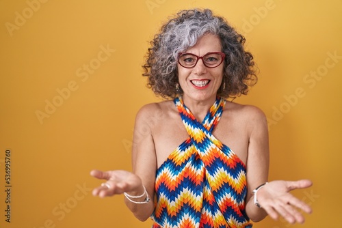 Middle age woman with grey hair standing over yellow background smiling cheerful offering hands giving assistance and acceptance.