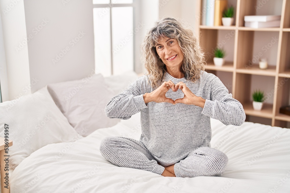 Middle age woman doing heart symbol with hands sitting on bed at bedroom Middle age woman doing heart symbol with hands sitting on bed at bedroom