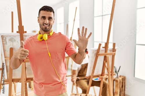 Young hispanic man at art studio showing and pointing up with fingers number six while smiling confident and happy.