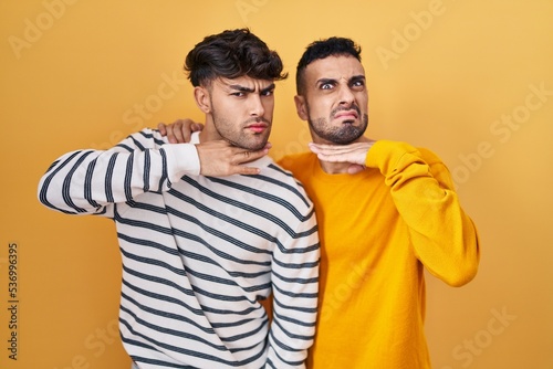 Young hispanic gay couple standing over yellow background cutting throat with hand as knife  threaten aggression with furious violence