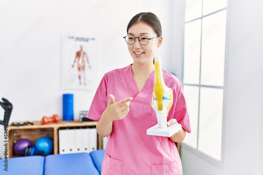 Young asian woman holding anatomical knee model at physiotherapy clinic