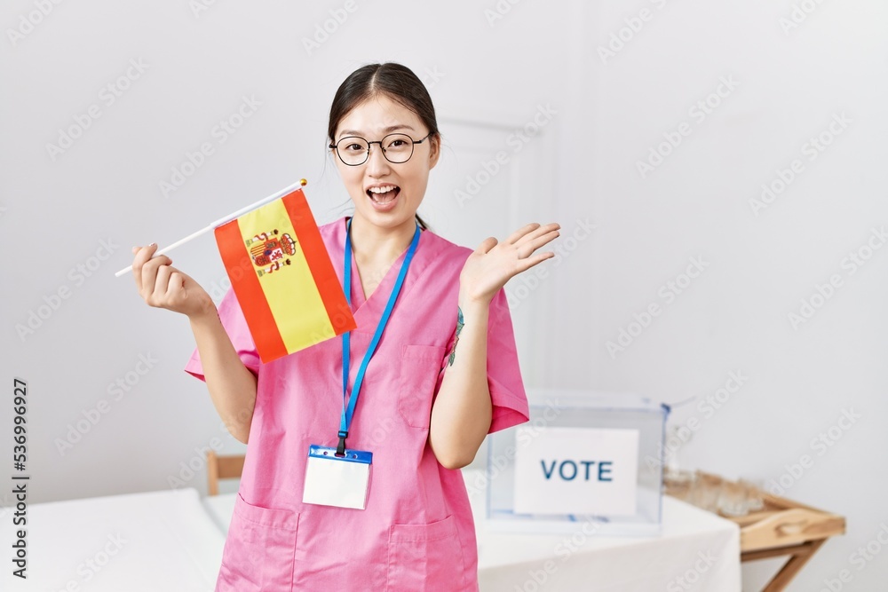 Young asian nurse woman at political campaign election holding spain flag celebrating achievement with happy smile and winner expression with raised hand Young asian nurse woman at political campaign election holding spain flag celebrating achievement with happy smile and winner expression with raised hand